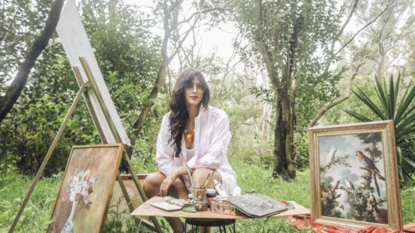 Woman artist seated outdoors with paints and brushes at a table, surrounded by framed artworks on the ground and easel art in progress.