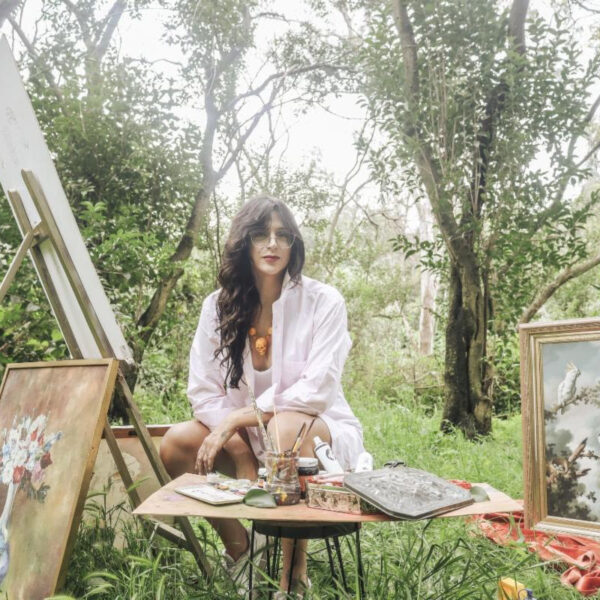 Woman artist seated outdoors with paints and brushes at a table, surrounded by framed artworks on the ground and easel art in progress.