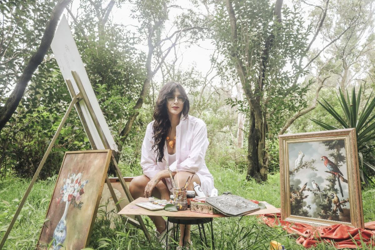 Woman artist seated outdoors with paints and brushes at a table, surrounded by framed artworks on the ground and easel art in progress.