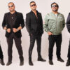 Five men wearing sunglasses pose in a studio, dressed in black jackets and casual outfits, lined up side by side.