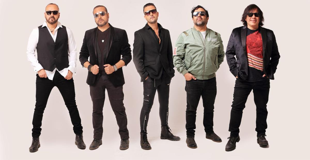 Five men wearing sunglasses pose in a studio, dressed in black jackets and casual outfits, lined up side by side.