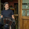 Man sits in doorway playing a black accordion outside a wooden storefront cafe.