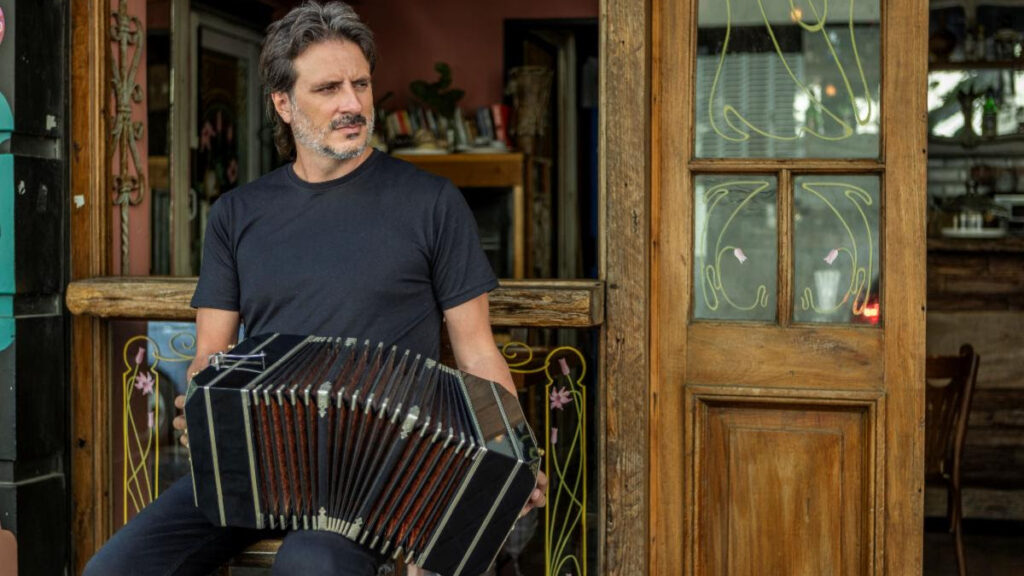 Man sits in doorway playing a black accordion outside a wooden storefront cafe.
