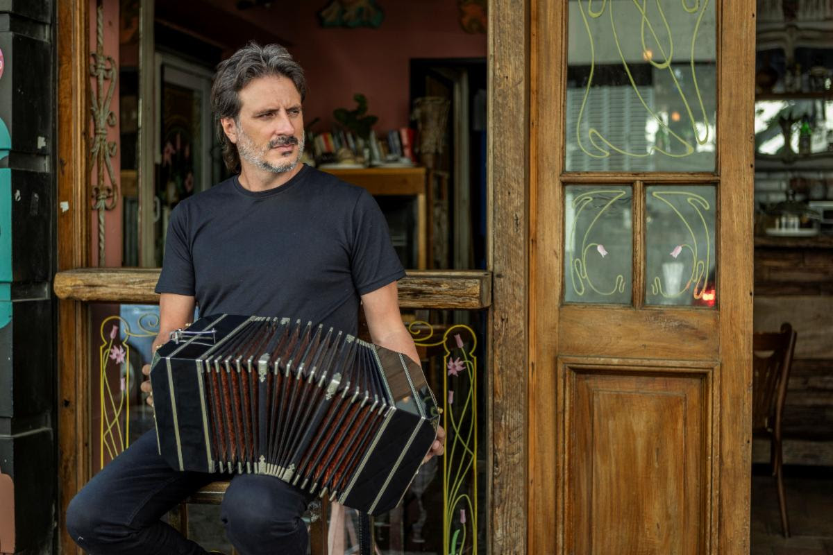 Man sits in doorway playing a black accordion outside a wooden storefront cafe.