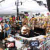 Live band performing in a busy music store, with saxophonist, guitarist, pianist, drummer, and a singer at center, surrounded by shelves and cameras.
