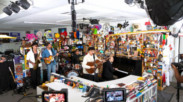 Live band performing in a busy music store, with saxophonist, guitarist, pianist, drummer, and a singer at center, surrounded by shelves and cameras.