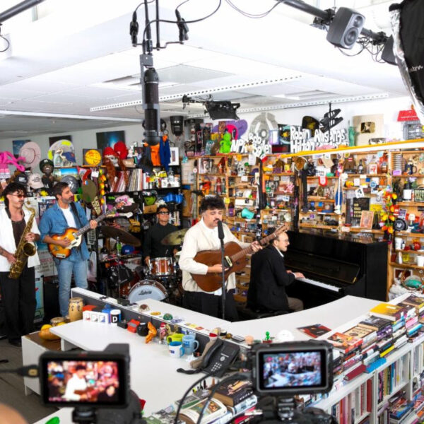 Live band performing in a busy music store, with saxophonist, guitarist, pianist, drummer, and a singer at center, surrounded by shelves and cameras.
