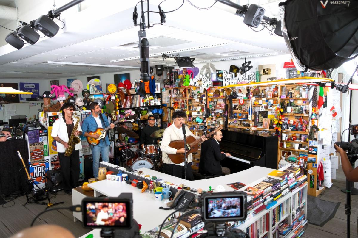 Live band performing in a busy music store, with saxophonist, guitarist, pianist, drummer, and a singer at center, surrounded by shelves and cameras.