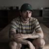 Bearded man in a striped shirt and green cap, crouching on a carpet and looking at the camera with arms resting on his knees.
