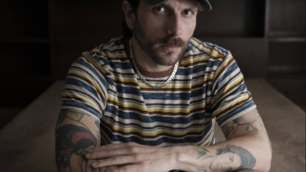 Bearded man in a striped shirt and green cap, crouching on a carpet and looking at the camera with arms resting on his knees.