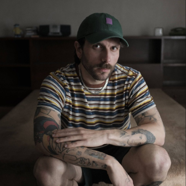 Bearded man in a striped shirt and green cap, crouching on a carpet and looking at the camera with arms resting on his knees.