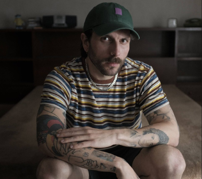 Bearded man in a striped shirt and green cap, crouching on a carpet and looking at the camera with arms resting on his knees.