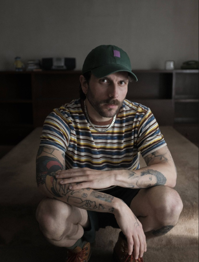 Bearded man in a striped shirt and green cap, crouching on a carpet and looking at the camera with arms resting on his knees.