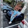 Man in blue shirt and light jeans standing beside a blue sports car in a parking lot, with a brick building in the background