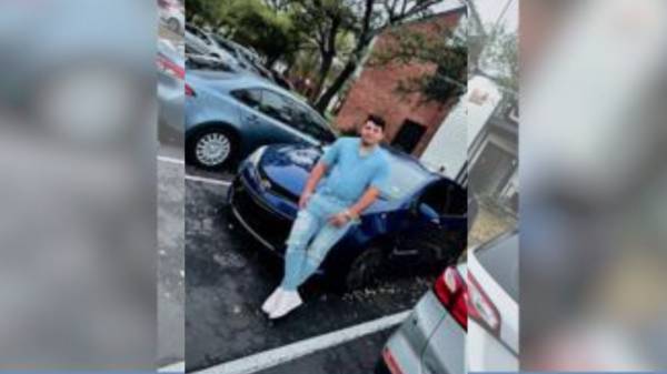 Man in blue shirt and light jeans standing beside a blue sports car in a parking lot, with a brick building in the background