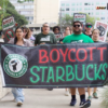 Protesters march holding a large banner that says 'BOYCOTT STARBUCKS' with a fist logo, in an urban street setting.