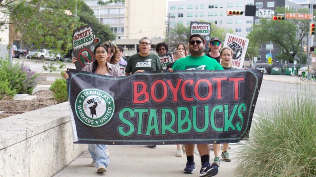 Protesters march holding a large banner that says 'BOYCOTT STARBUCKS' with a fist logo, in an urban street setting.
