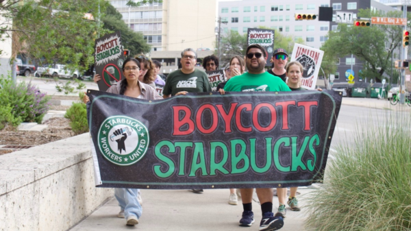 Protesters march holding a large banner that says 'BOYCOTT STARBUCKS' with a fist logo, in an urban street setting.