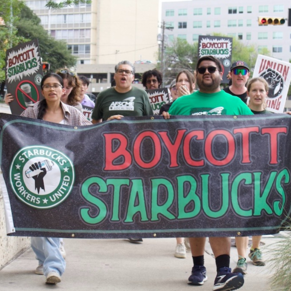 Protesters march holding a large banner that says 'BOYCOTT STARBUCKS' with a fist logo, in an urban street setting.