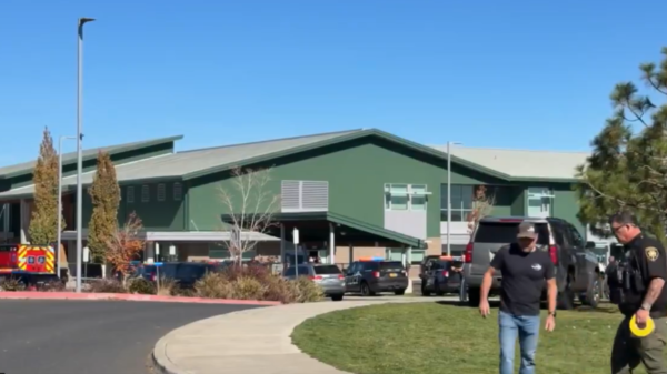 Green building with a sloped roof and parked cars, emergency responders visible outside on a sunny day.