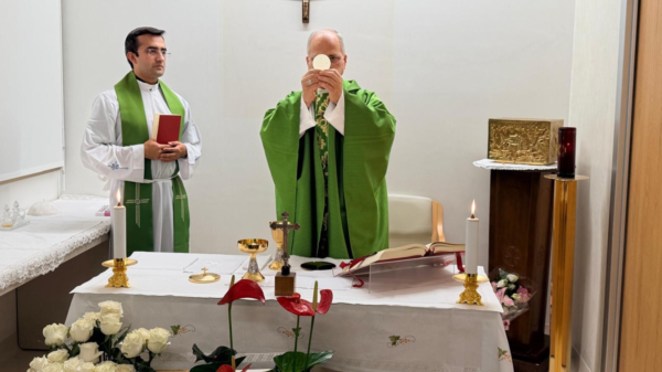 Two priests in green vestments stand at an altar during a Catholic Mass, one presenting the Eucharist as the other looks on, with candles and sacred vessels nearby