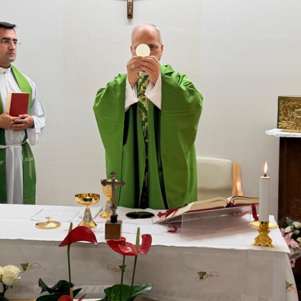 Two priests in green vestments stand at an altar during a Catholic Mass, one presenting the Eucharist as the other looks on, with candles and sacred vessels nearby