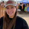 Smiling woman wearing a Texas cap at an outdoor market with colorful clothing stalls in the background.