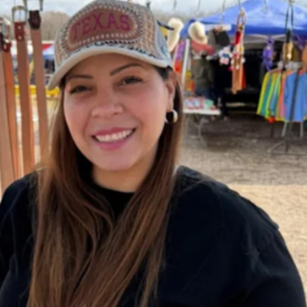 Smiling woman wearing a Texas cap at an outdoor market with colorful clothing stalls in the background.