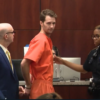 A man in an orange jumpsuit being escorted by a police officer toward a desk in a courtroom.