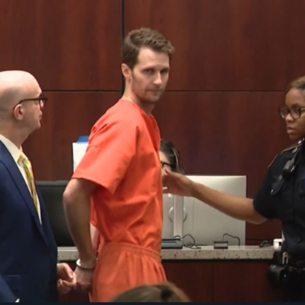 A man in an orange jumpsuit being escorted by a police officer toward a desk in a courtroom.