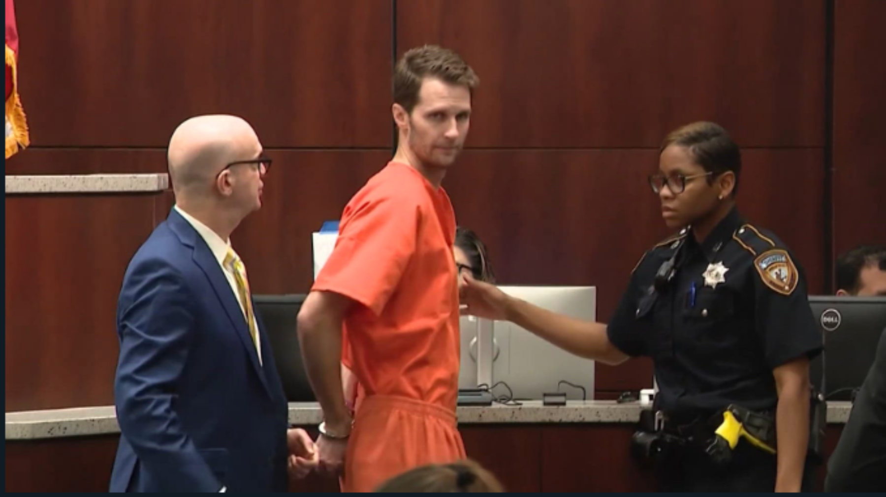 A man in an orange jumpsuit being escorted by a police officer toward a desk in a courtroom.