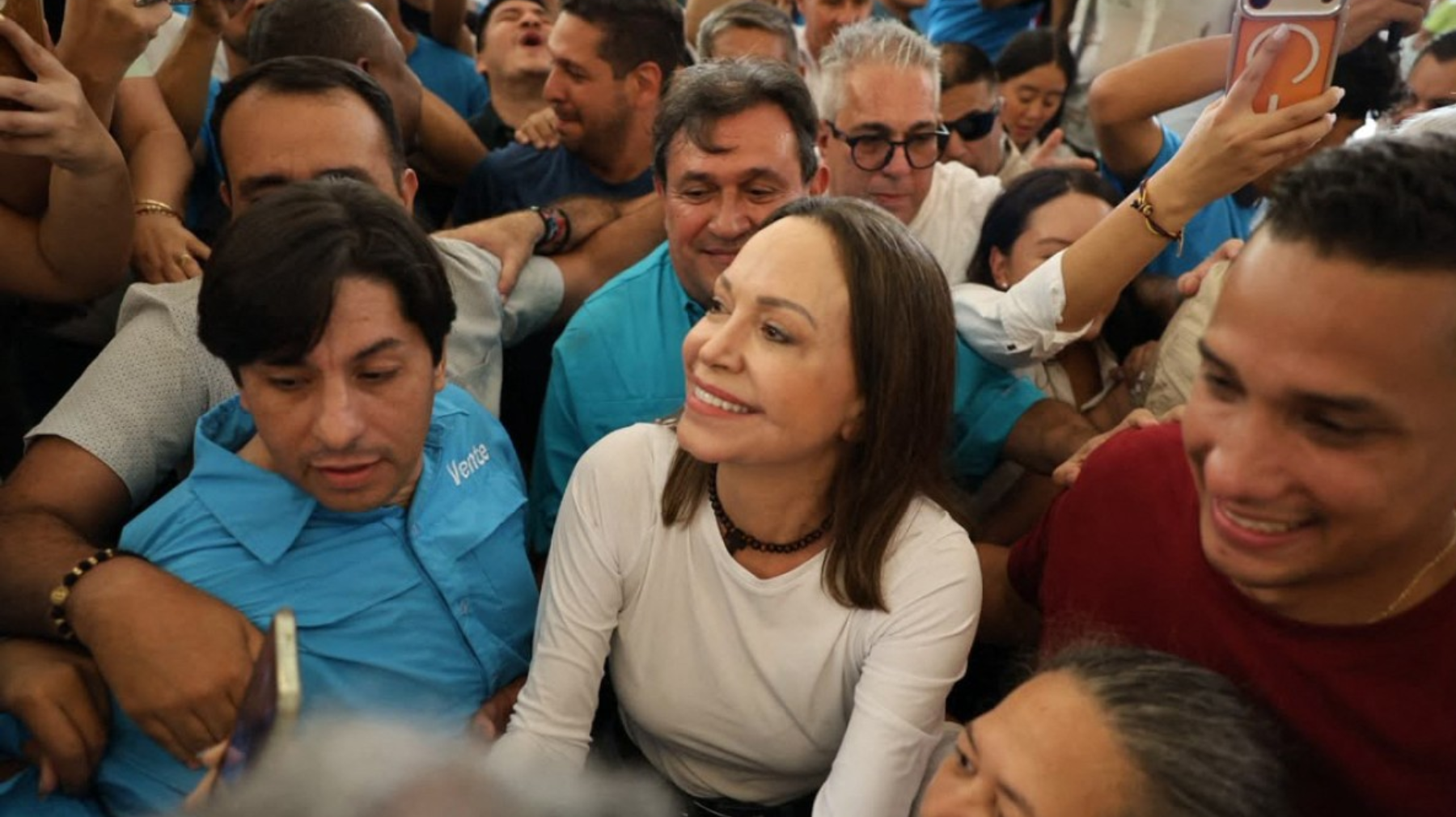Smiling woman in a white shirt sits among a dense crowd taking selfies at a public event.