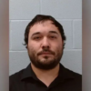 Head-and-shoulders mugshot of a man with dark hair and a beard, wearing a black shirt, against a gray cinder block wall.