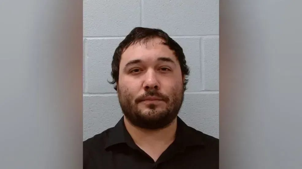 Head-and-shoulders mugshot of a man with dark hair and a beard, wearing a black shirt, against a gray cinder block wall.