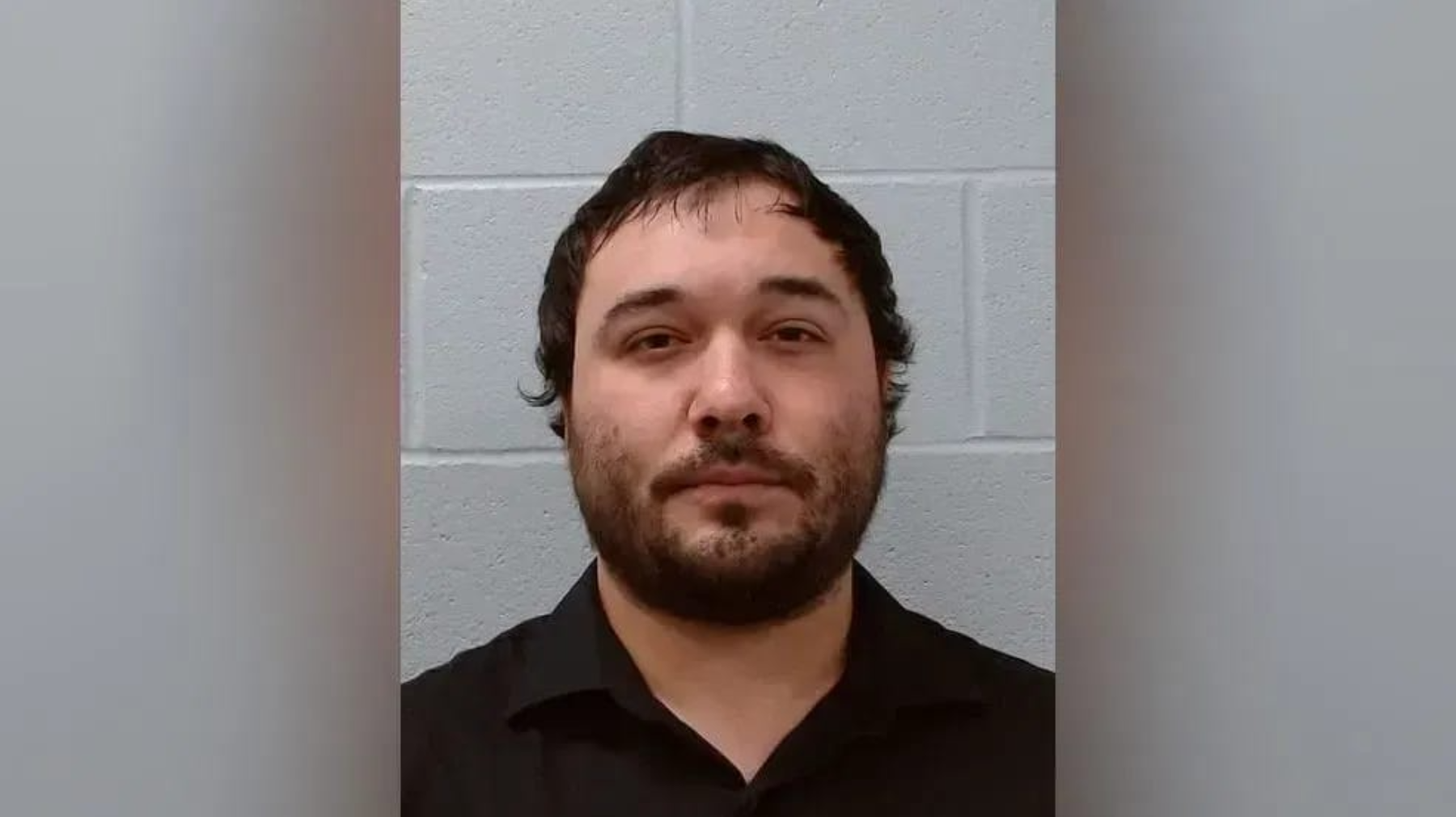 Head-and-shoulders mugshot of a man with dark hair and a beard, wearing a black shirt, against a gray cinder block wall.