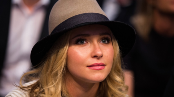 Close-up of a young woman with blonde hair wearing a beige fedora with a black band, looking upward.