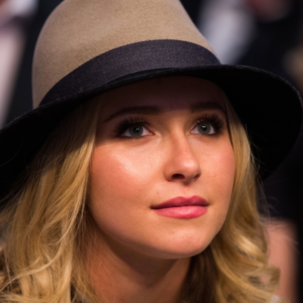 Close-up of a young woman with blonde hair wearing a beige fedora with a black band, looking upward.