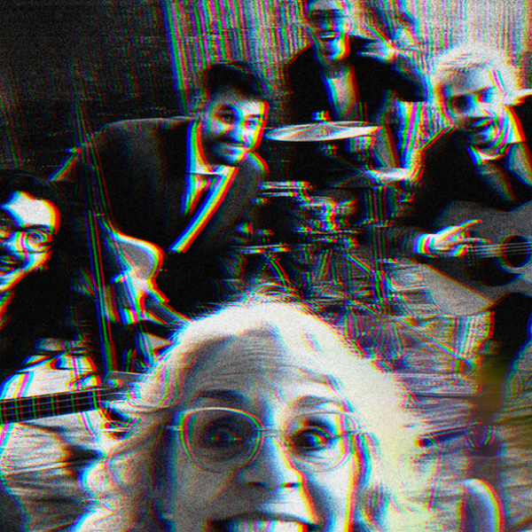 Older woman in front taking a selfie with a four‑piece band in a rehearsal space, features guitar, violin, and drums; image has a red/blue glitch effect.
