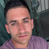 Close-up selfie of a man with short dark hair wearing a mauve shirt, looking at the camera.
