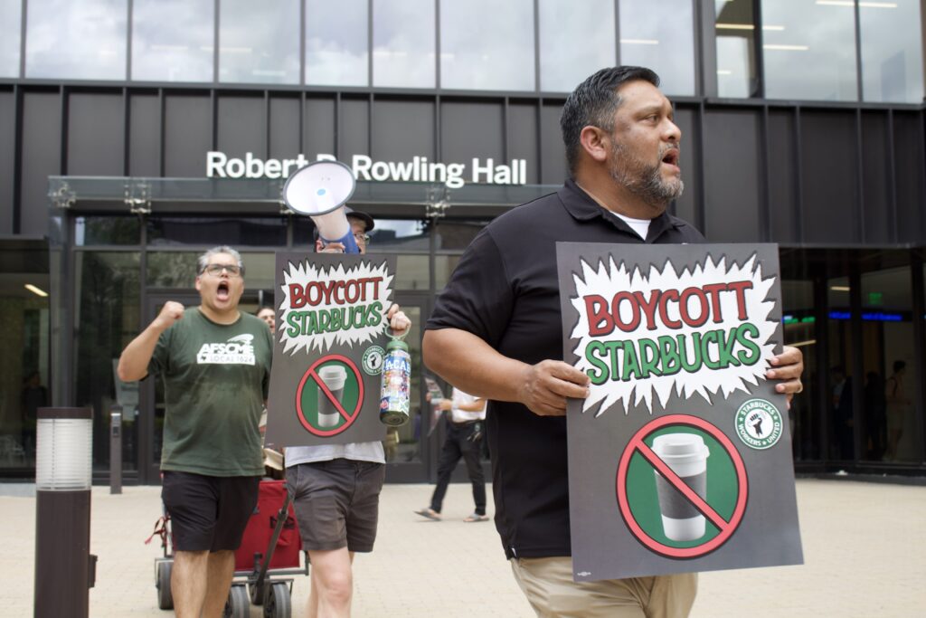 Group of protesters outside a modern building holding 'Boycott Starbucks' signs; a man in a black polo leads the scene.