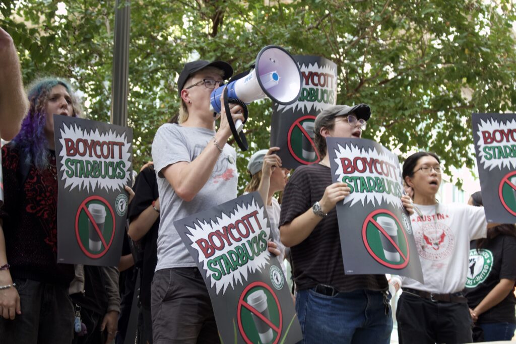 Group of protesters holding signs reading'Boycott Starbucks' with a person using a megaphone in the center. Trees in the background.