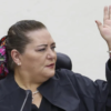 Woman raising her right hand while speaking, wearing a dark blazer and colorful scarf tied in her hair, bracelets on the wrist.