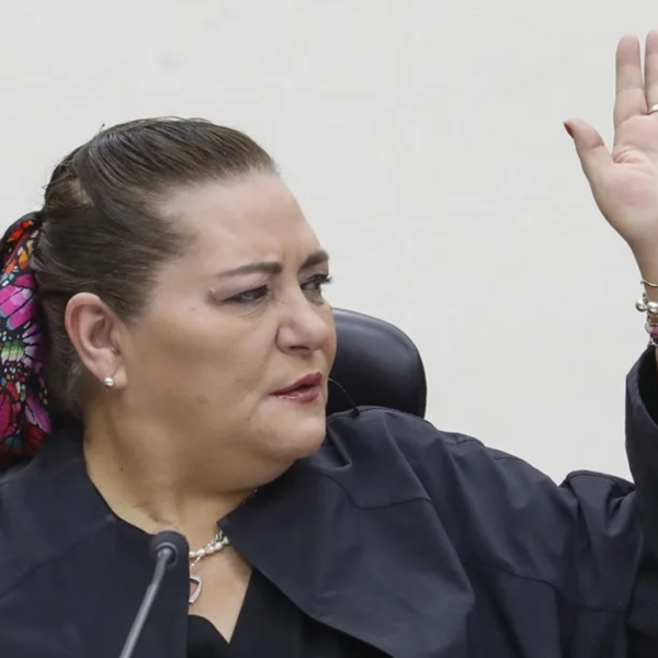 Woman raising her right hand while speaking, wearing a dark blazer and colorful scarf tied in her hair, bracelets on the wrist.