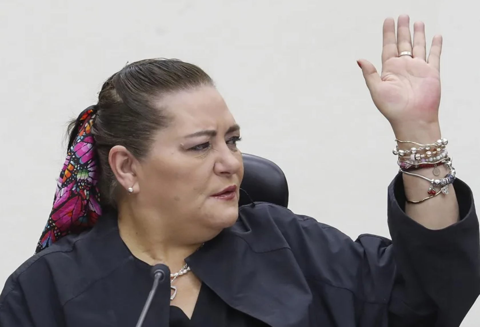 Woman raising her right hand while speaking, wearing a dark blazer and colorful scarf tied in her hair, bracelets on the wrist.