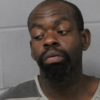 Mugshot of a man with a goatee wearing a gray striped jail uniform, looking to his left against a gray wall.