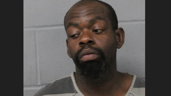 Mugshot of a man with a goatee wearing a gray striped jail uniform, looking to his left against a gray wall.