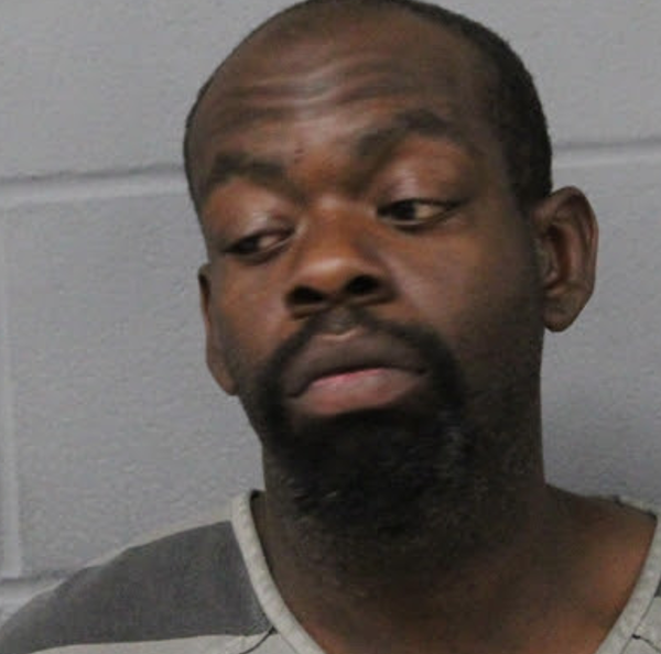 Mugshot of a man with a goatee wearing a gray striped jail uniform, looking to his left against a gray wall.