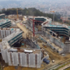 Aerial view of a large construction site with multiple connected concrete buildings, cranes, and construction equipment across a cleared site near a city edge.