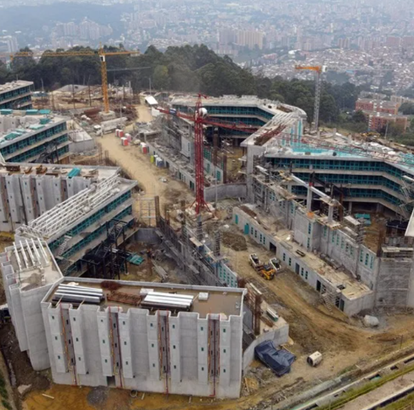 Aerial view of a large construction site with multiple connected concrete buildings, cranes, and construction equipment across a cleared site near a city edge.