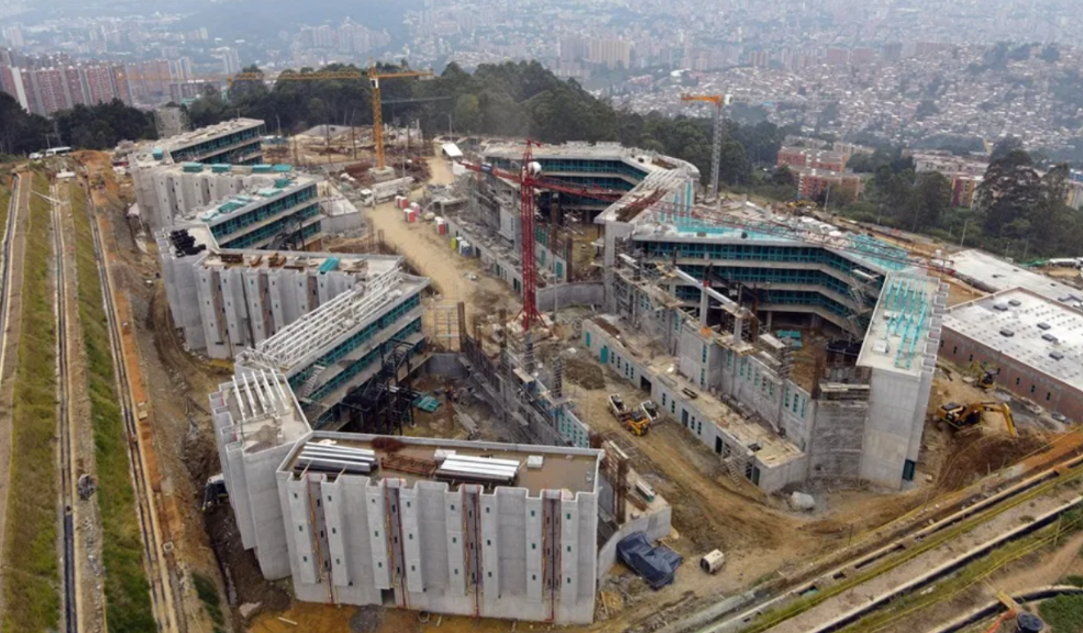 Aerial view of a large construction site with multiple connected concrete buildings, cranes, and construction equipment across a cleared site near a city edge.
